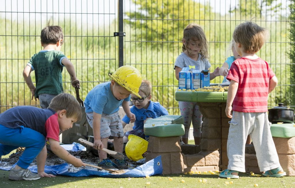 A group of small children , some in yellow hard hats, play in the mud near a playground picnic table