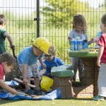 A group of small children , some in yellow hard hats, play in the mud near a playground picnic table