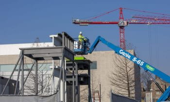 Construction worker in a cherry picker places a top element on a metal structure with a red crane in the background