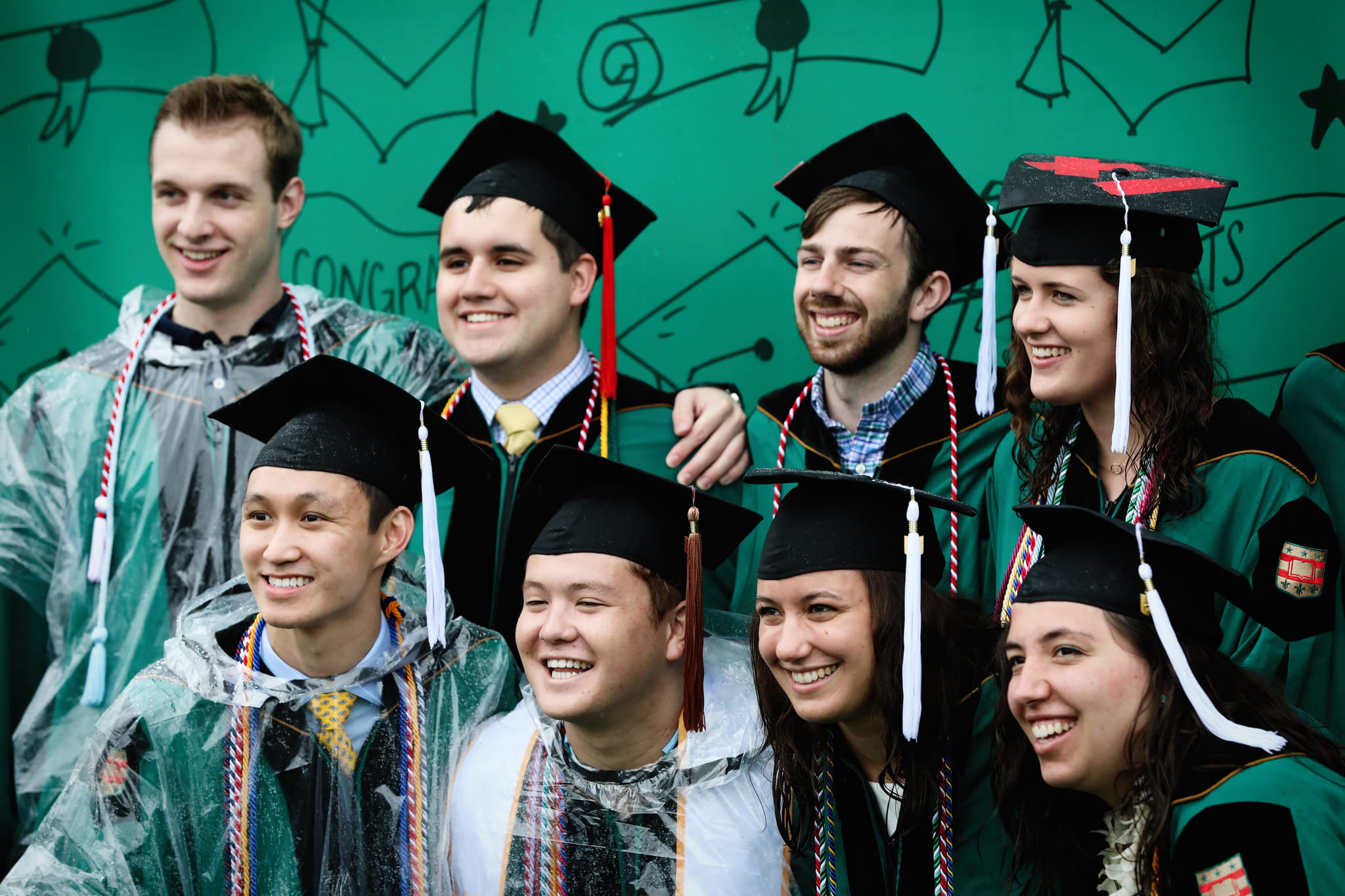 A group of WashU students in cap and gown pose for photos in front of a green illustrated backdrop